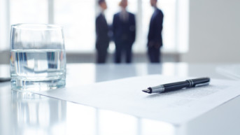 Image of business document, pen and glass of water at workplace with group of colleagues on background